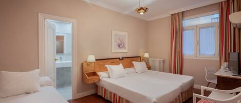 A hotel room with two beds, a wooden headboard, a desk with a chair, a radiator, and a window with curtains.