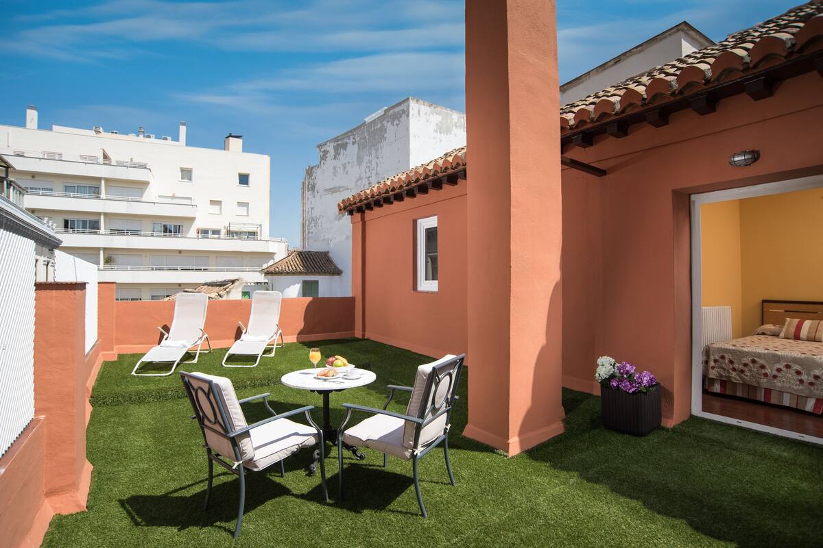 A rooftop terrace with artificial grass, two lounge chairs, a small table with fruit, and a potted plant.