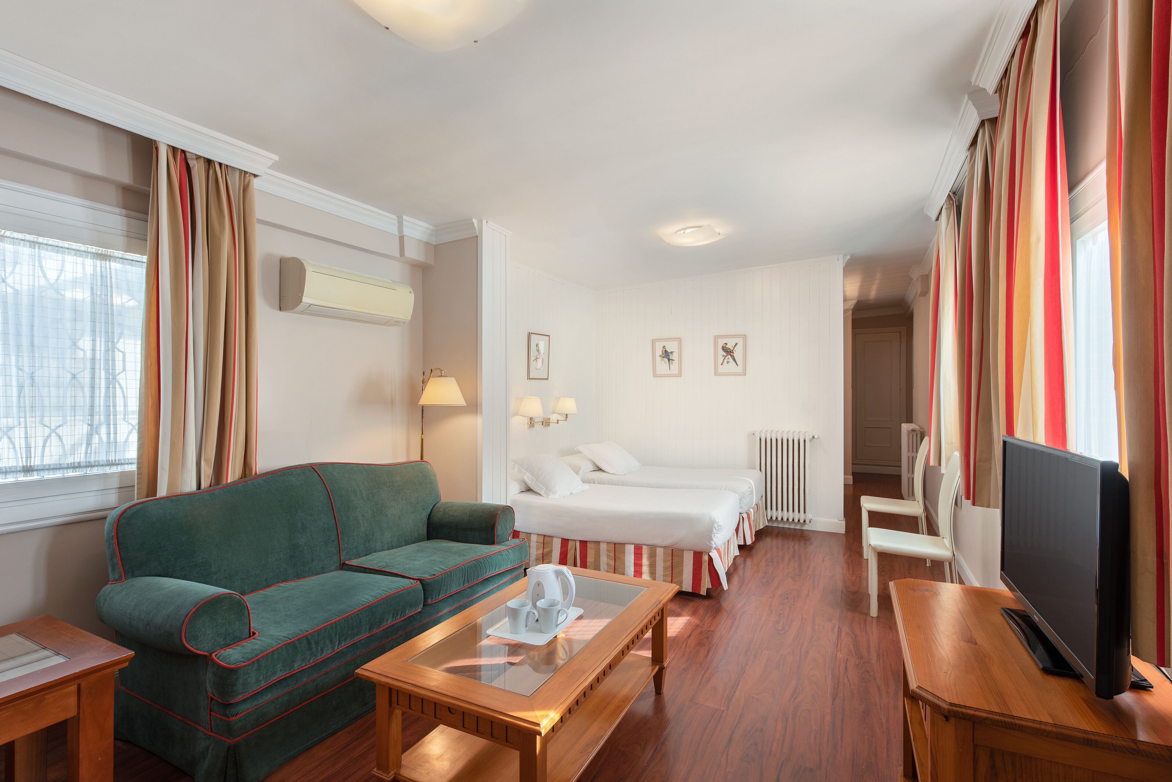 Ein Hotelzimmer mit einem grünen Sofa, einem Holztisch, einem Fernseher, einem Bett mit weißer Bettwäsche und einem Fenster mit Vorhängen.