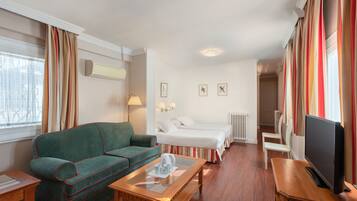 Una habitación de hotel con un sofá verde, una mesa de centro de madera, un televisor, una cama con ropa blanca y un ventanal con cortinas.