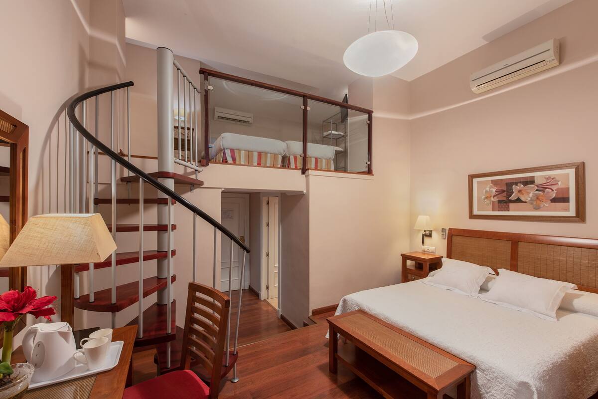 A hotel room with a bed, a desk, a chair, a small table, a lamp, a picture frame, a mirror, a staircase, and an air conditioning unit.