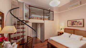 Habitación de hotel con cama, escritorio, silla, pequeña mesa, lámpara, marco de fotos, espejo, escalera y unidad de aire acondicionado.