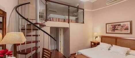 A hotel room with a bed, a desk, a chair, a small table, a lamp, a picture frame, a mirror, a staircase, and an air conditioning unit.