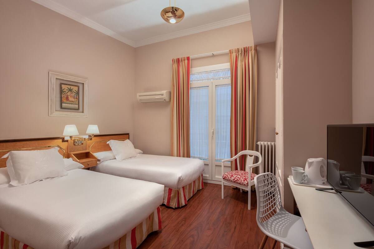 A hotel room with two beds, a desk, a chair, a TV, and a window with curtains.