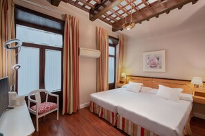A hotel room with a wooden ceiling, a bed with white linens, a chair with a red and white cushion, and a desk with a television and a lamp.