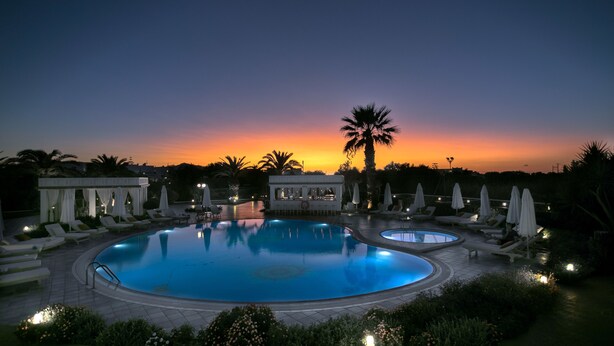 Zona de piscina con sillones de descanso y sombrillas, rodeada de vegetación y un atardecer de fondo.