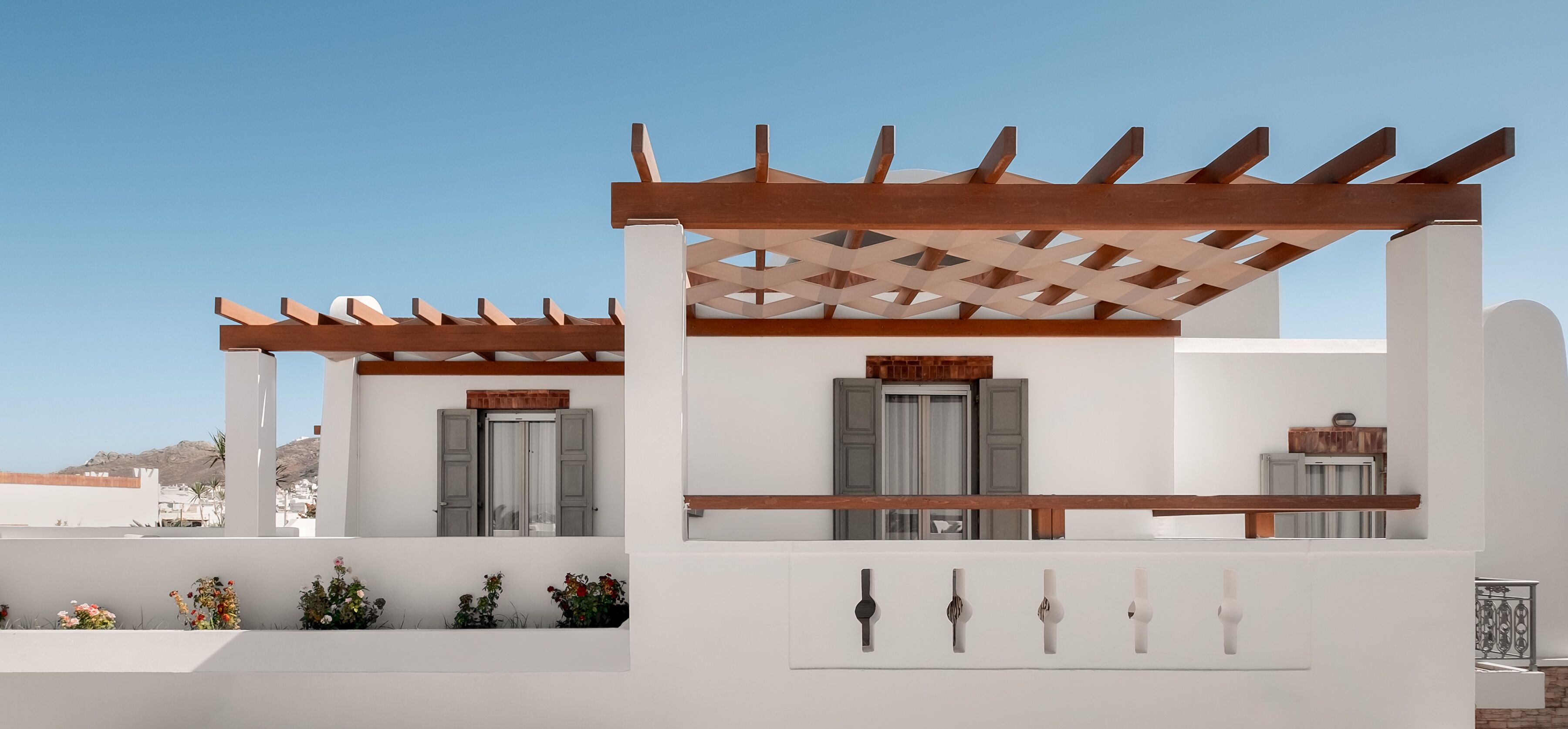 A white building with a wooden pergola, grey shutters, and a small balcony with potted plants.
