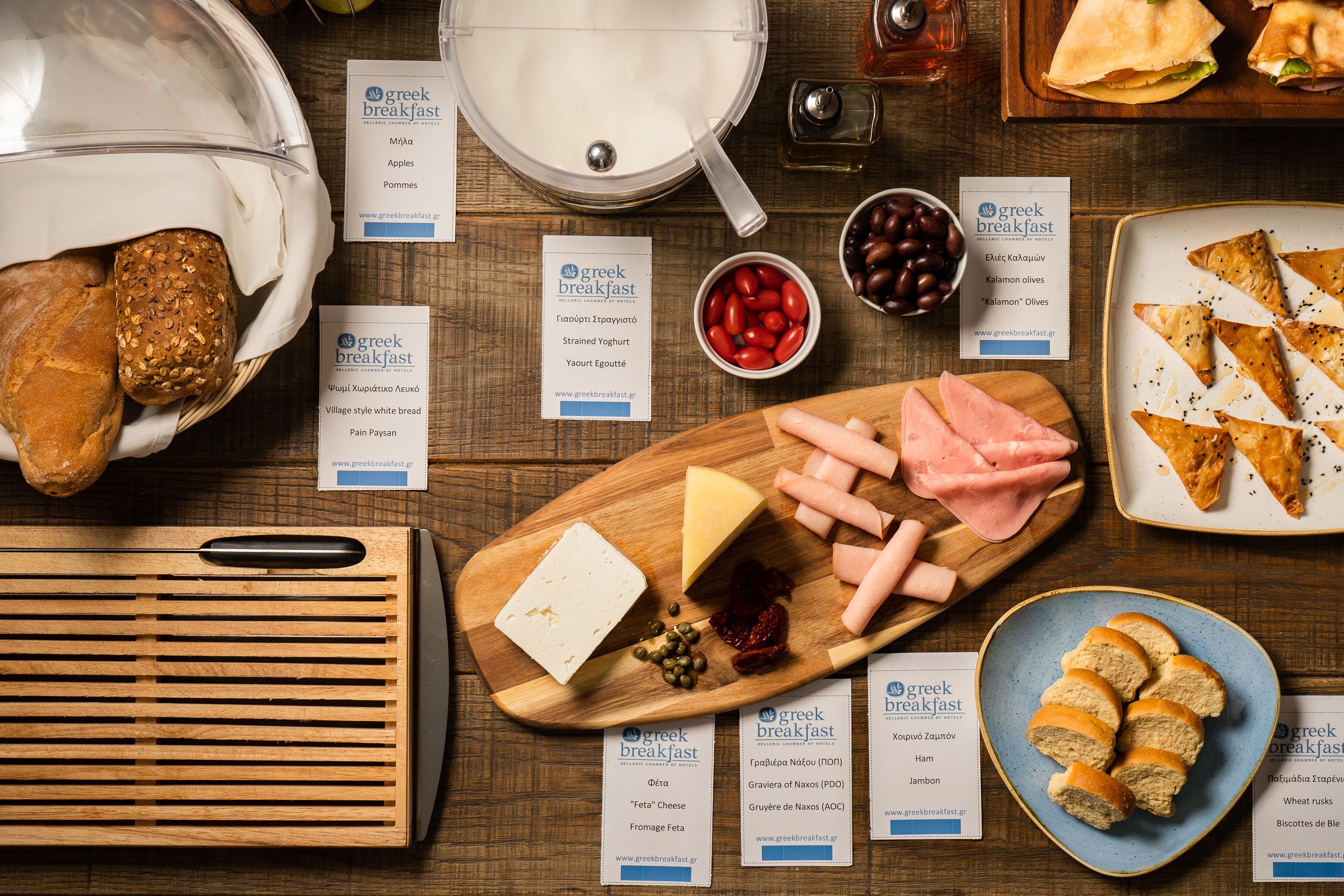 A wooden table with various food items and Greek breakfast cards.