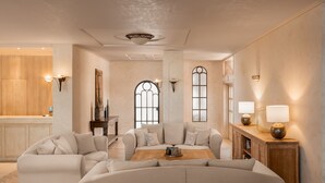 Amplio salón con un gran sofá en forma de L, una mesa de centro de madera y un aparador de madera con lámparas. Hay dos ventanas grandes con vidrios decorativos y una puerta de madera con marco de metal.