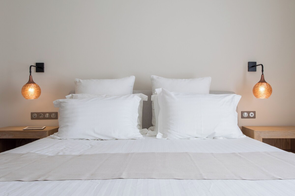 A neatly made bed with white bedding and pillows, flanked by wooden nightstands with wall-mounted lamps.