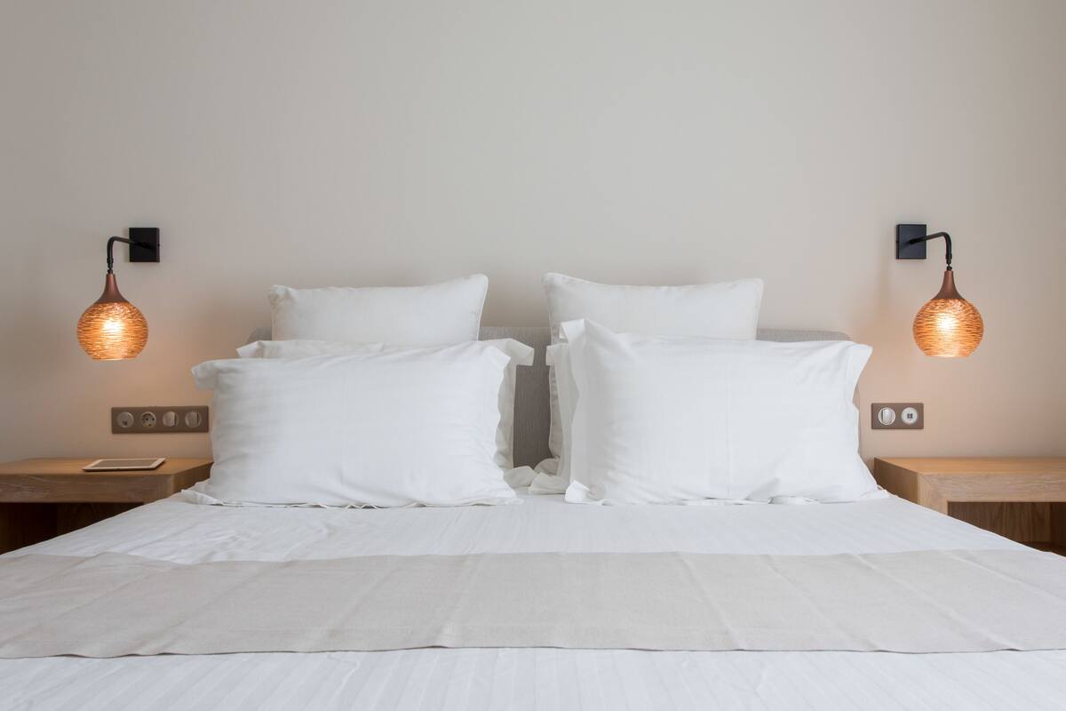 A neatly made bed with white linens and pillows, two bedside tables with lamps, and a wall-mounted light fixture.