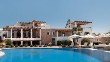 A large swimming pool with sun loungers and umbrellas, surrounded by a building with multiple balconies and a stone facade.
