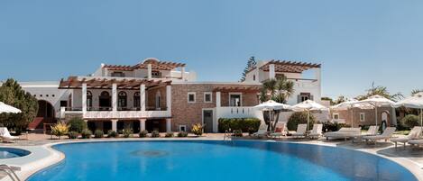 A large swimming pool with sun loungers and umbrellas, surrounded by a building with multiple balconies and a stone facade.