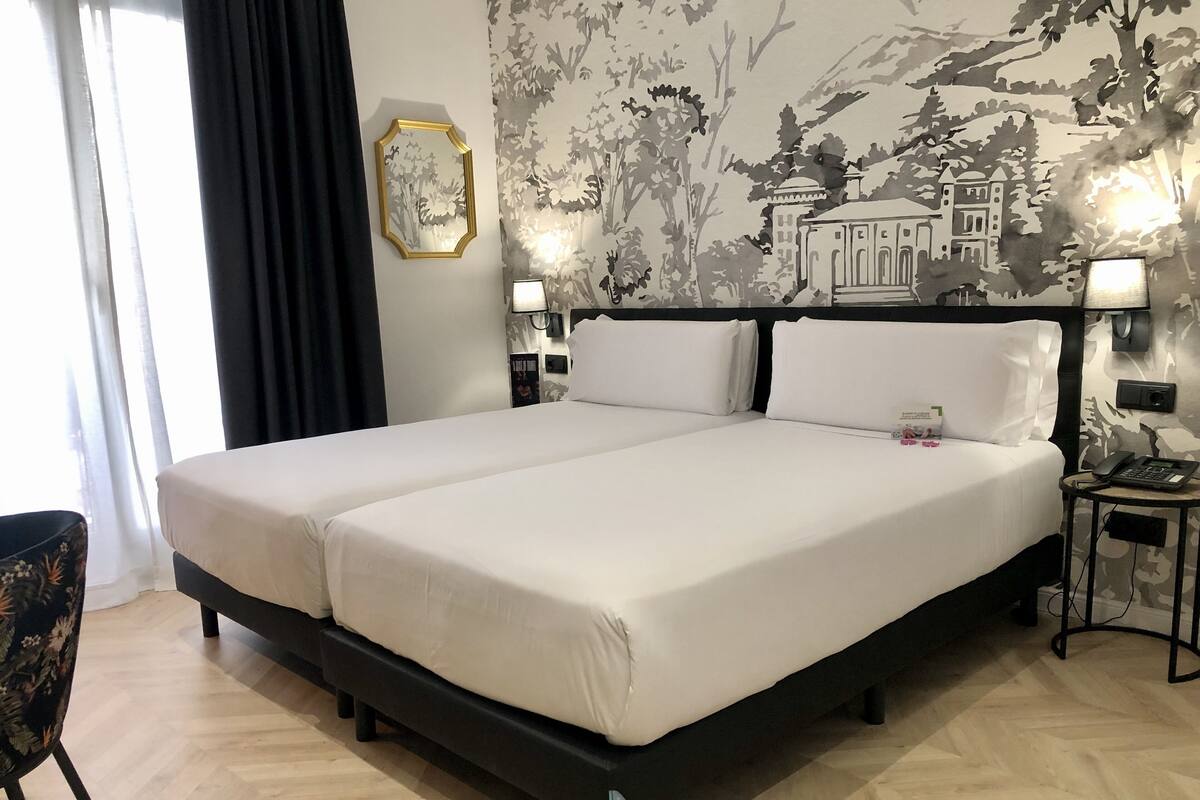 Una cama doble con ropa blanca, dos mesitas de noche con lámparas, un papel tapiz con estampado floral y una silla con diseño floral.