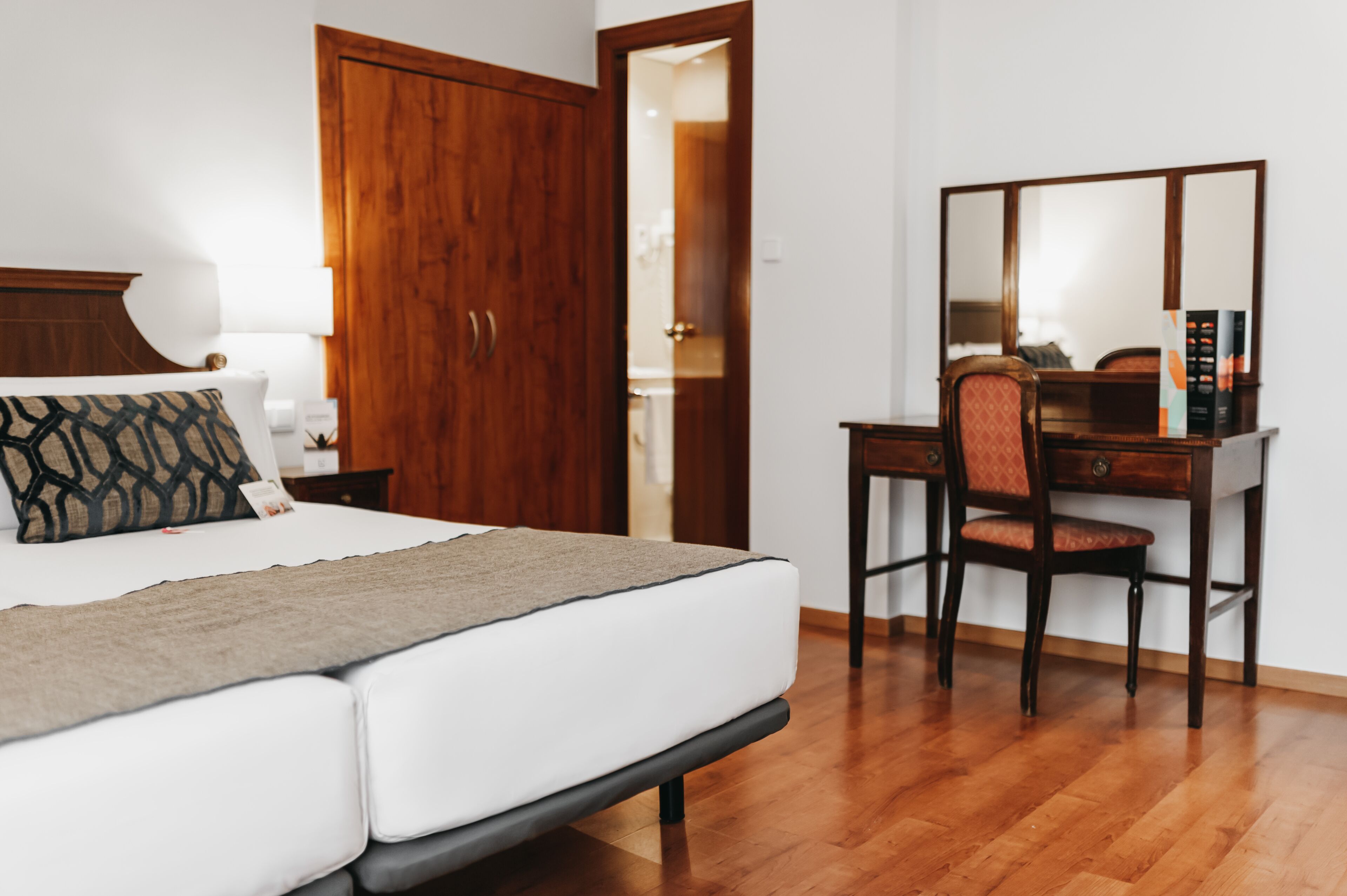 A bedroom with a bed, a wooden wardrobe, a desk with a chair, and a mirror.
