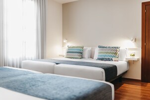 A hotel room with two beds, a bedside table with a lamp, a small shelf with a plant, and a window with curtains.