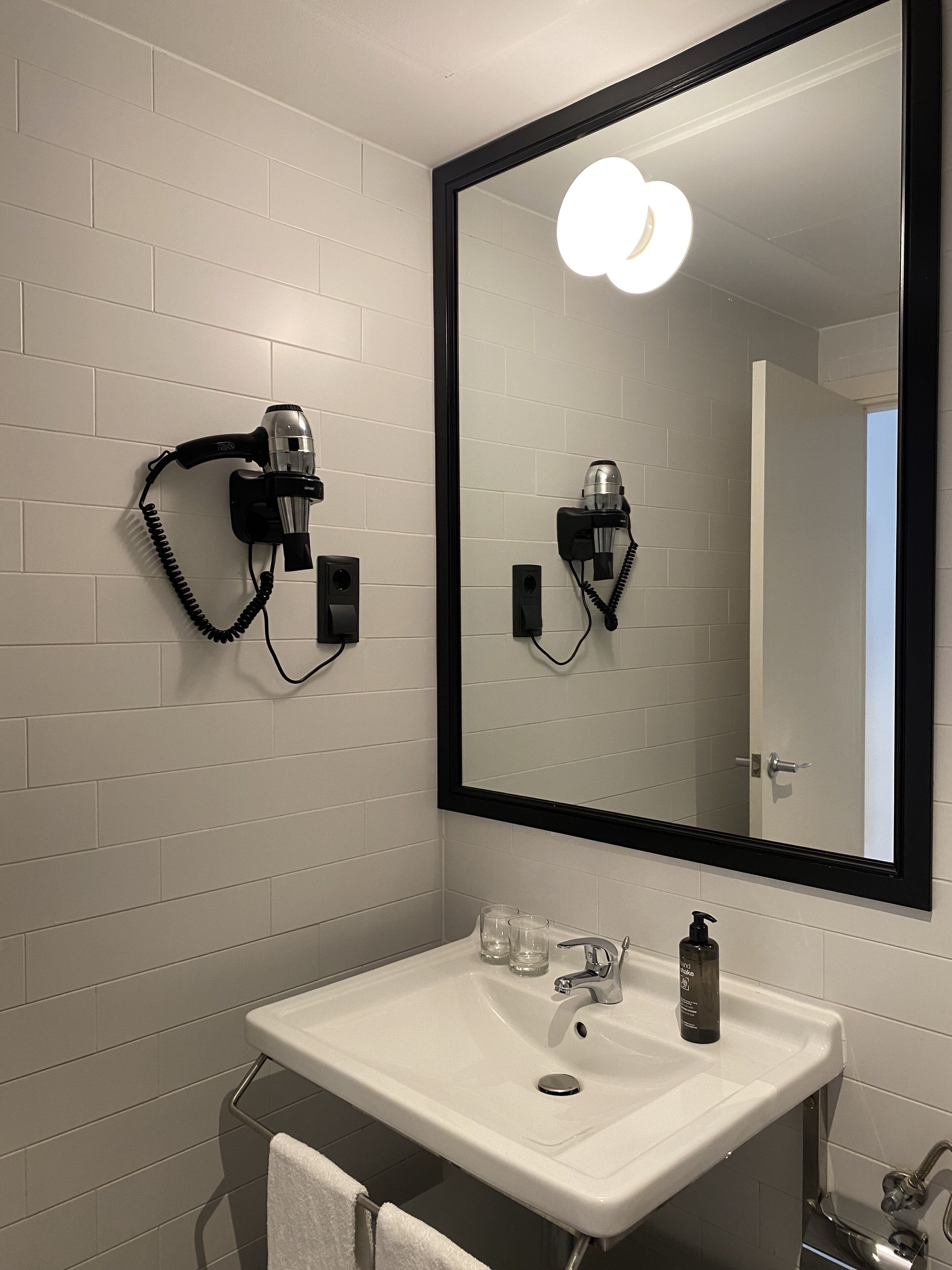 Un baño con un lavamanos blanco, dos secadores de pelo fijos en la pared, un espejo con marco negro y un perchero para toallas.