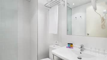 Un baño moderno con cabina de ducha de vidrio, inodoro blanco, lavabo con artículos de aseo y un secador de pelo montado en la pared.