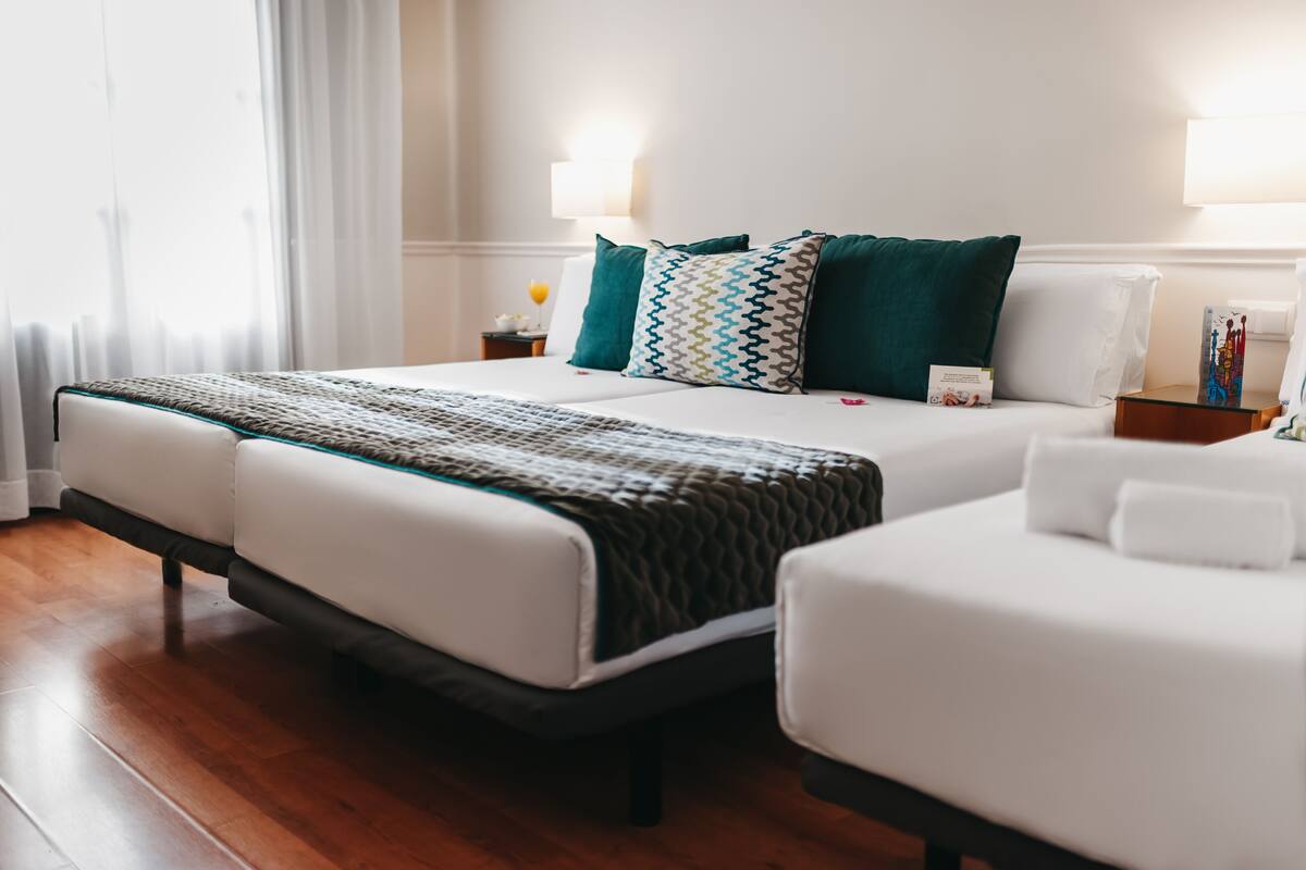 Habitación de hotel con dos camas, una mesita de noche, una lámpara y una ventana con cortinas.