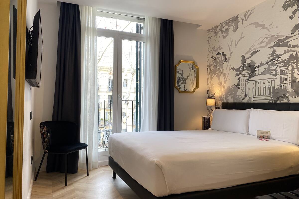 Habitación de hotel con cama, una silla, televisión, ventana con cortinas y un mural en la pared.
