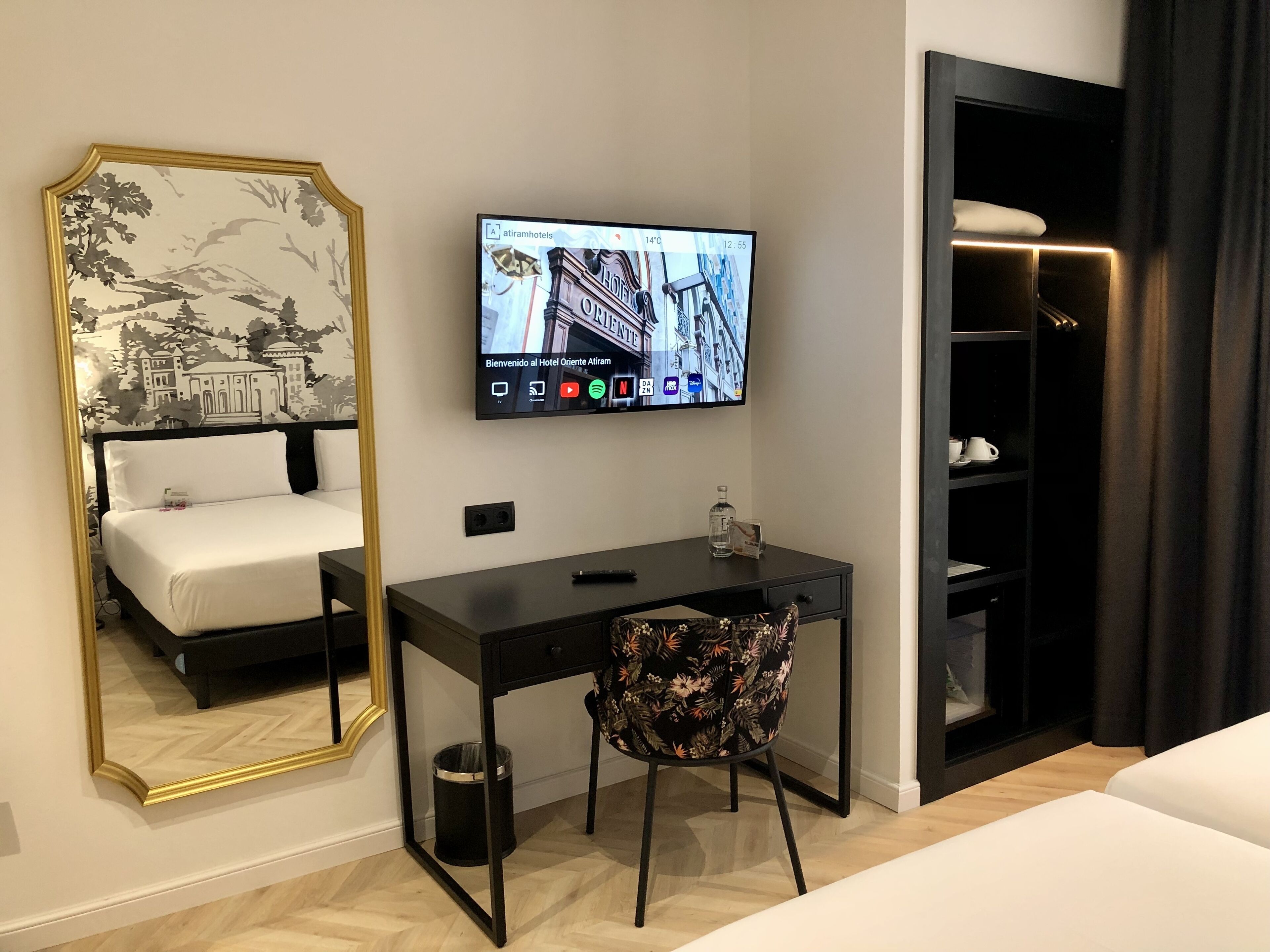 Una habitación de hotel con cama, escritorio, silla, televisor montado en la pared y armario.