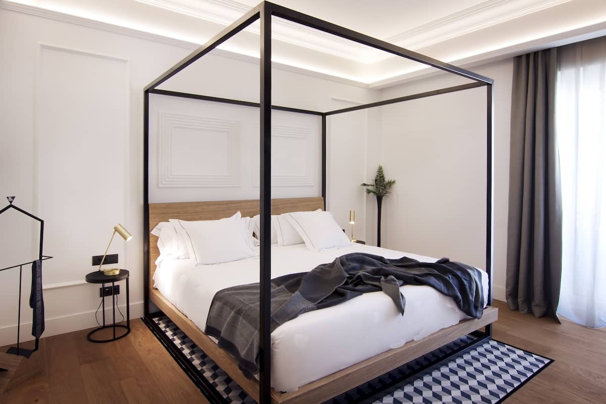A four-poster bed with a wooden frame and white bedding, a small side table with a lamp, and a checkered rug on a wooden floor.