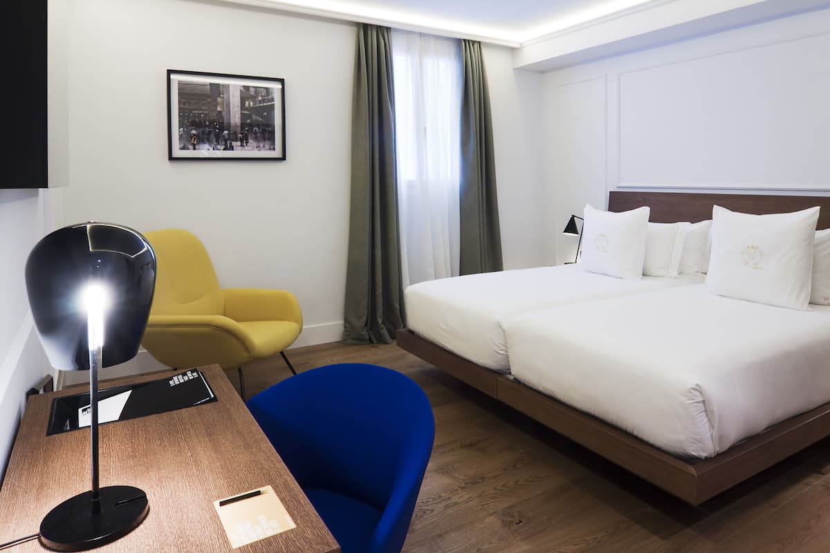 A hotel room with a bed, a desk with a lamp, a blue chair, a yellow chair, and a framed picture on the wall.