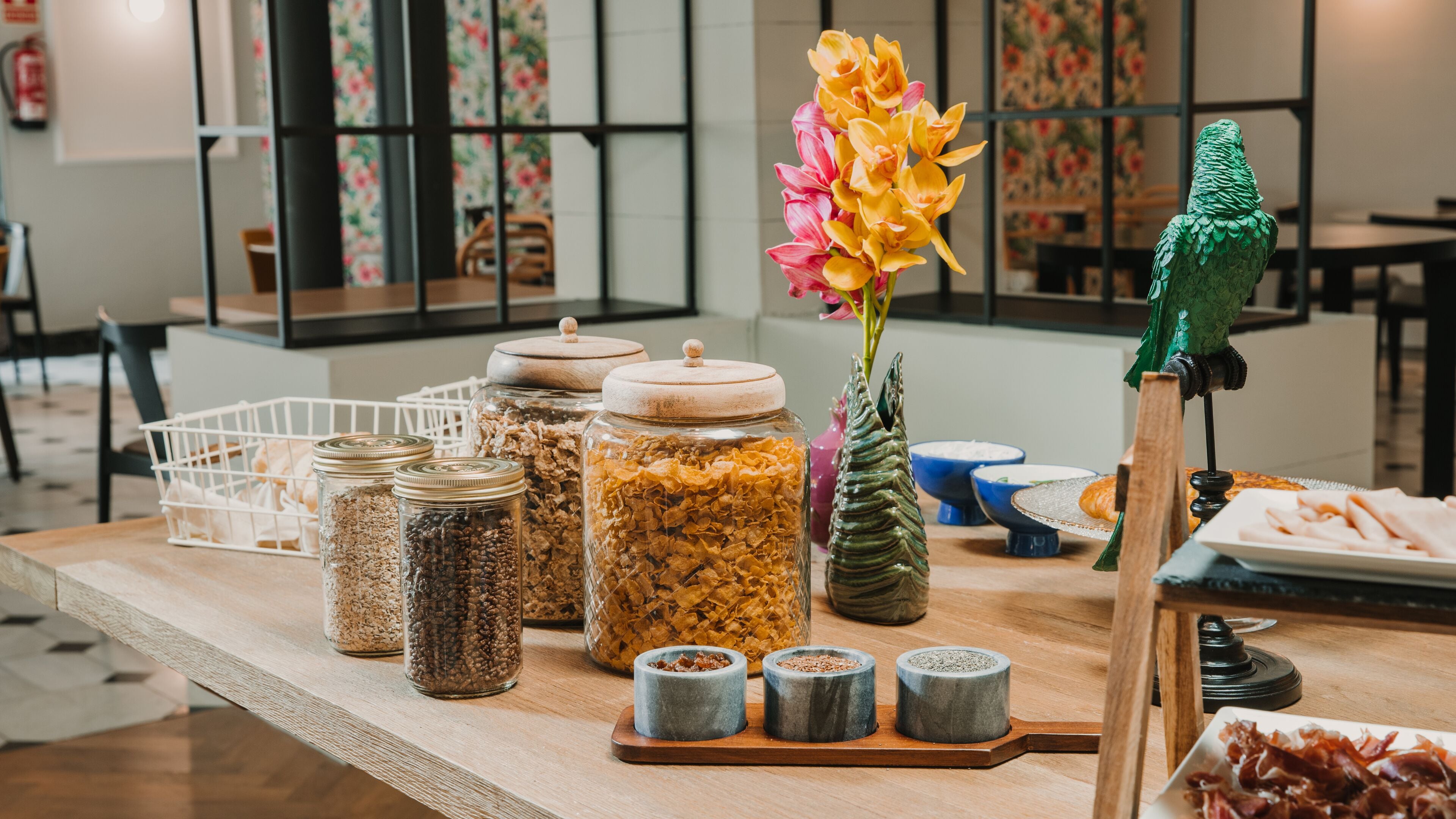 Una barra de desayuno con diversos alimentos, incluyendo botes de granola, un florero con flores y una figurita de loro.