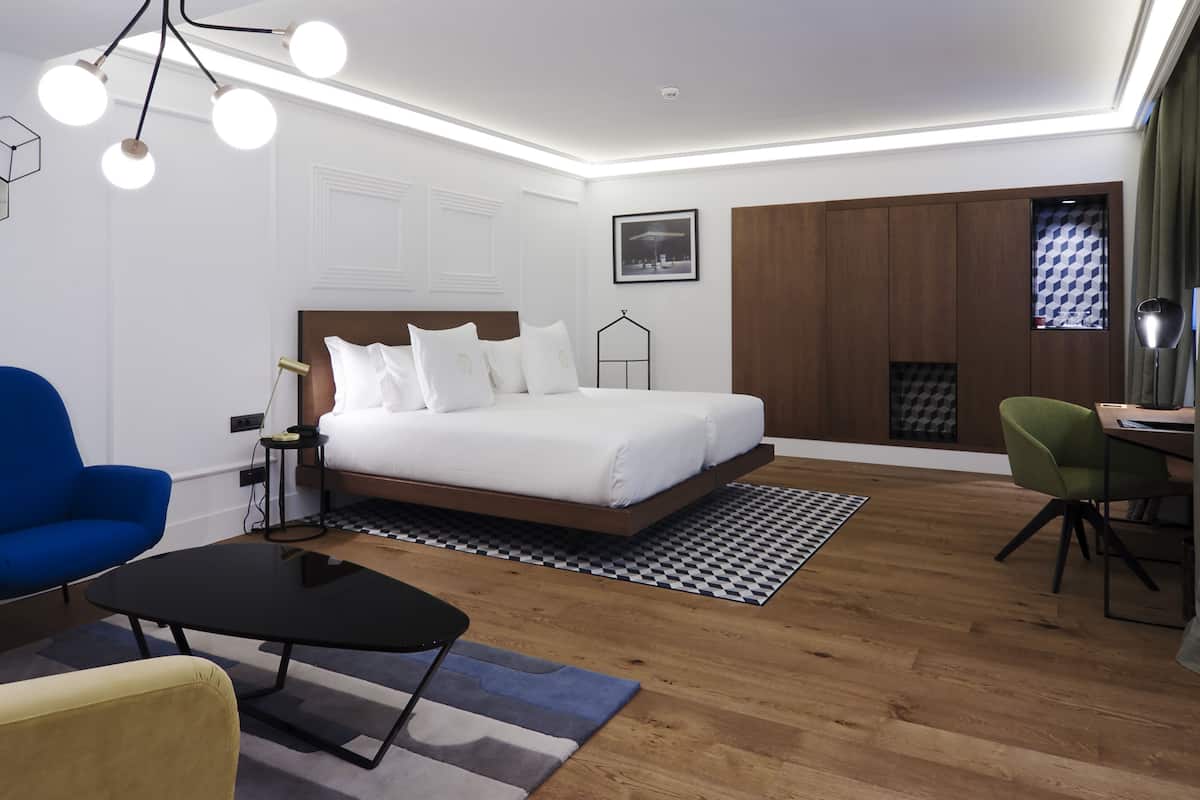 A modern hotel room with a large bed, a desk, a blue chair, a green chair, and a black coffee table.