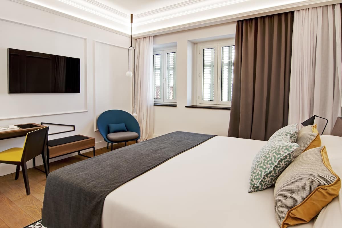 A hotel room with a bed, a desk, a chair, a TV mounted on the wall, and windows with curtains.