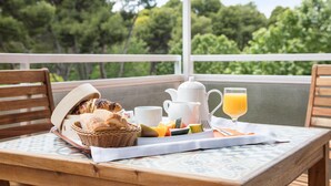 Un desayuno con pan, frutas, una tetera, una taza y un vaso de zumo de naranja sobre una mesa con vistas a árboles.