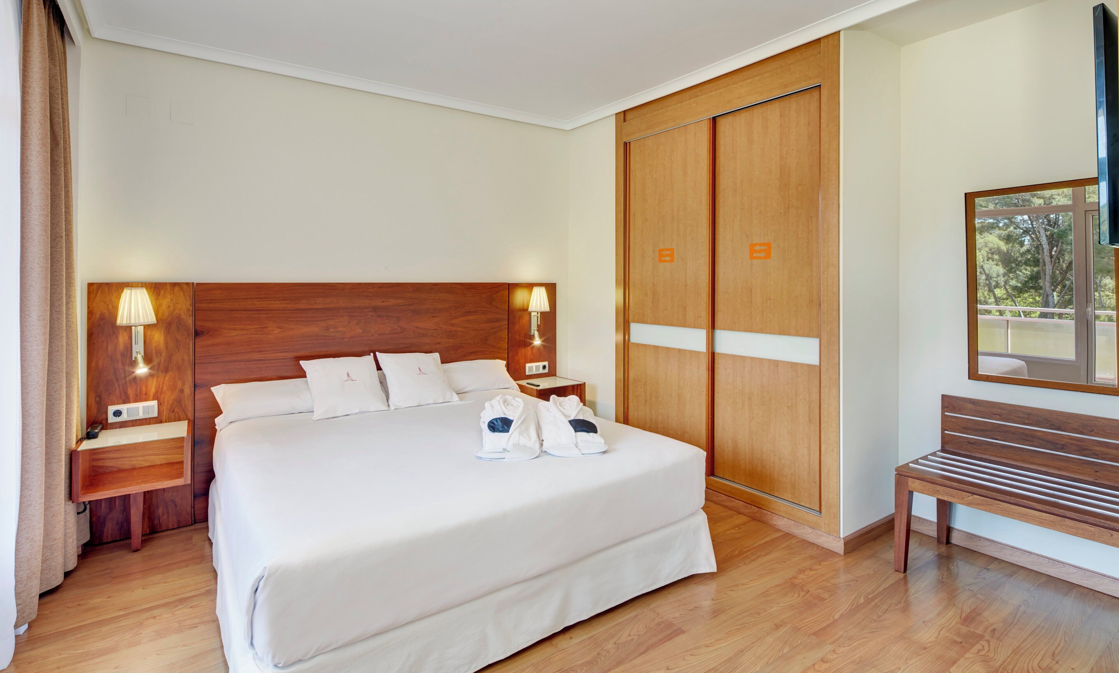 Una habitación de hotel con una cama grande, cabecero de madera, dos mesitas de noche con lámparas, un armario, un banco y un espejo grande.