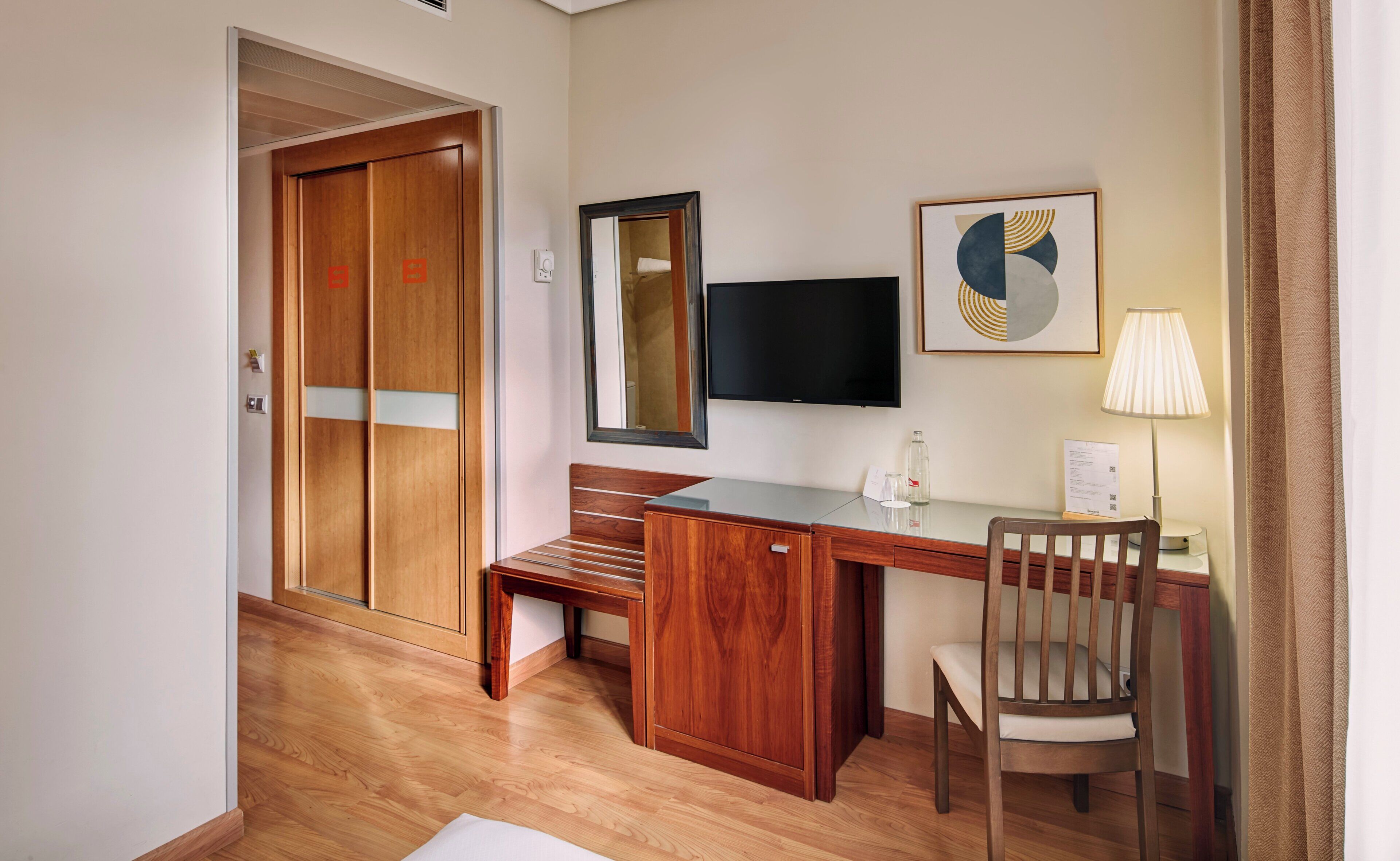 Habitación de hotel con un escritorio de madera, una silla, un televisor de pantalla plana, un cuadro abstracto enmarco y una lámpara.