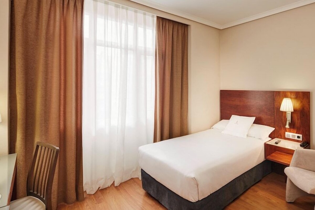 A hotel room with a bed, a nightstand with a lamp, a chair, and a desk with a chair.