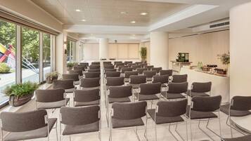 Un salón de conferencias con filas de sillas, una estación de café y grandes ventanales con vista a un parque infantil.