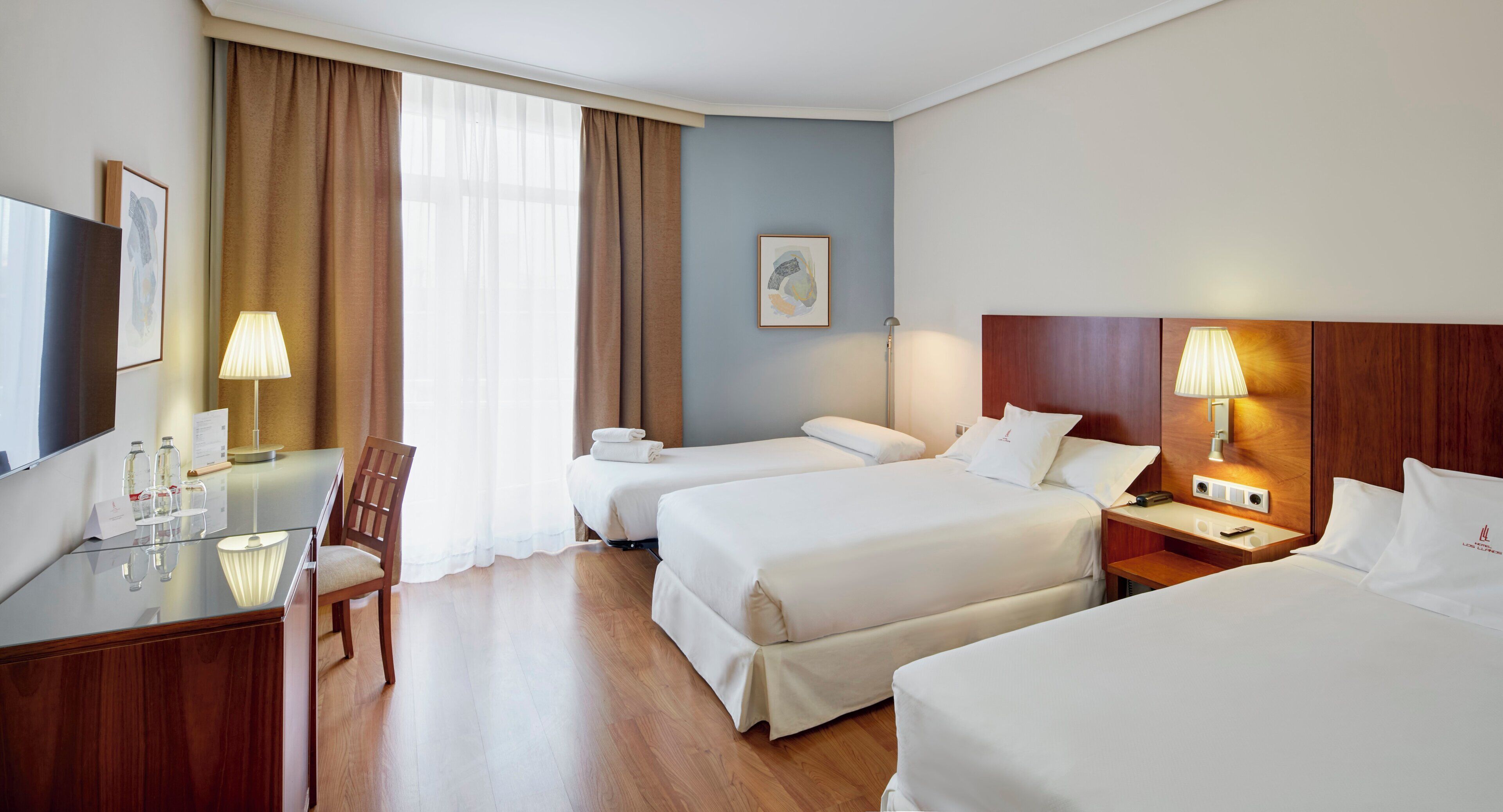 A hotel room with two beds, a desk, a chair, a TV, and a lamp.
