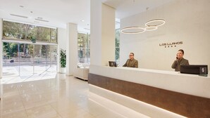 Zona de recepción de un hotel moderno con un diseño limpio y minimalista, que incluye un mostrador de recepción, un amplio ventanal con vista a la calle y un cartel que dice "LOS LLANOS".