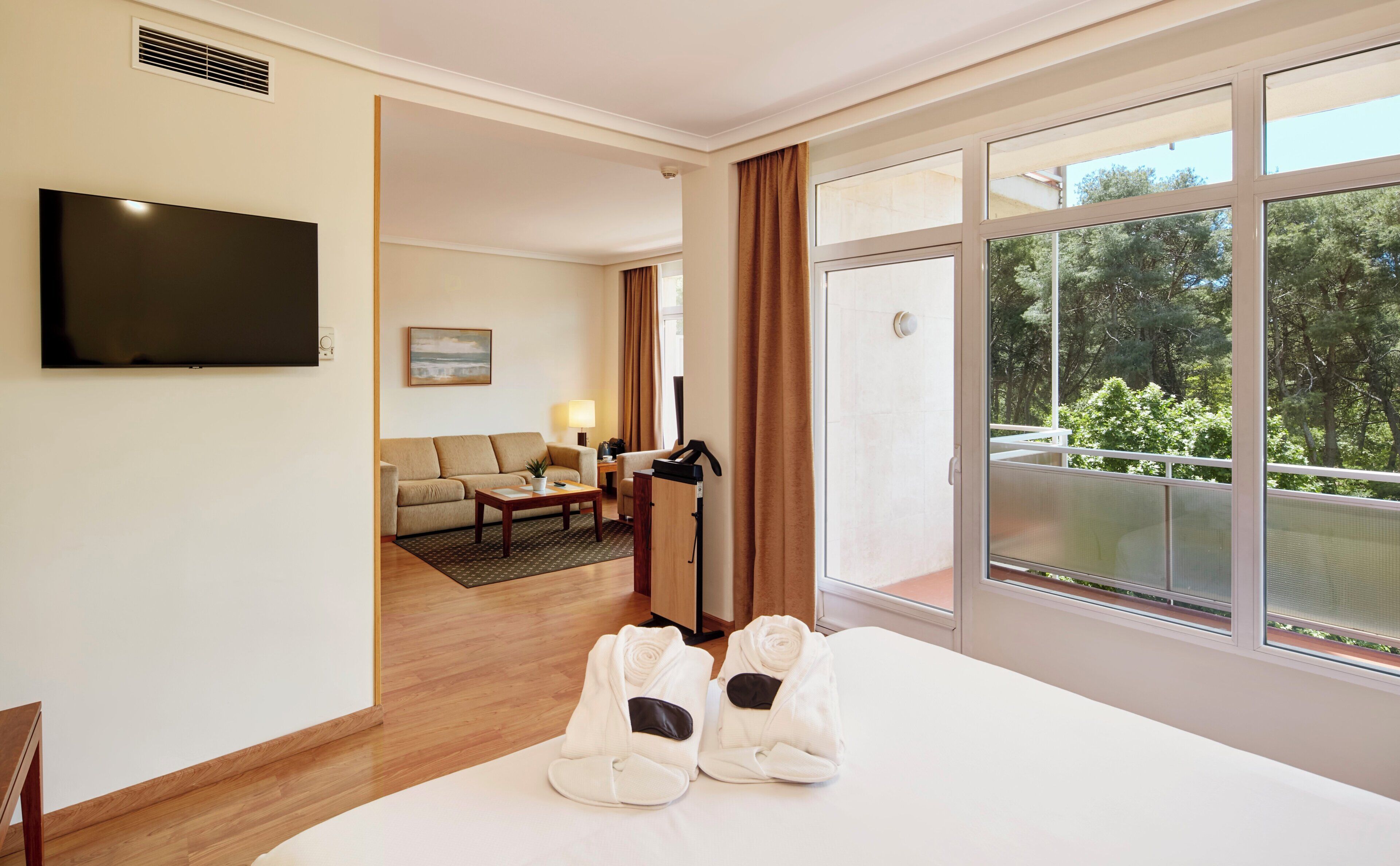 A hotel room with a large window, a flat-screen TV, a sofa, a coffee table, and a pair of slippers.