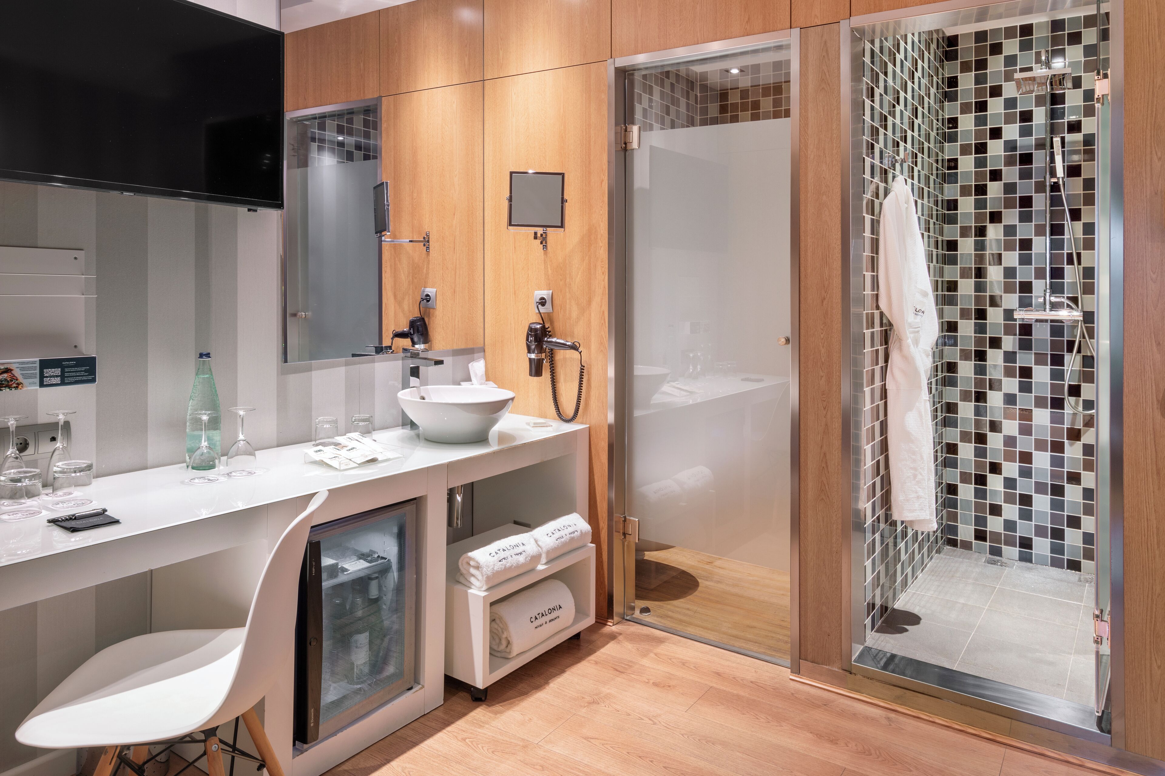 A modern bathroom with a vanity, a shower with a glass door, and a small seating area.