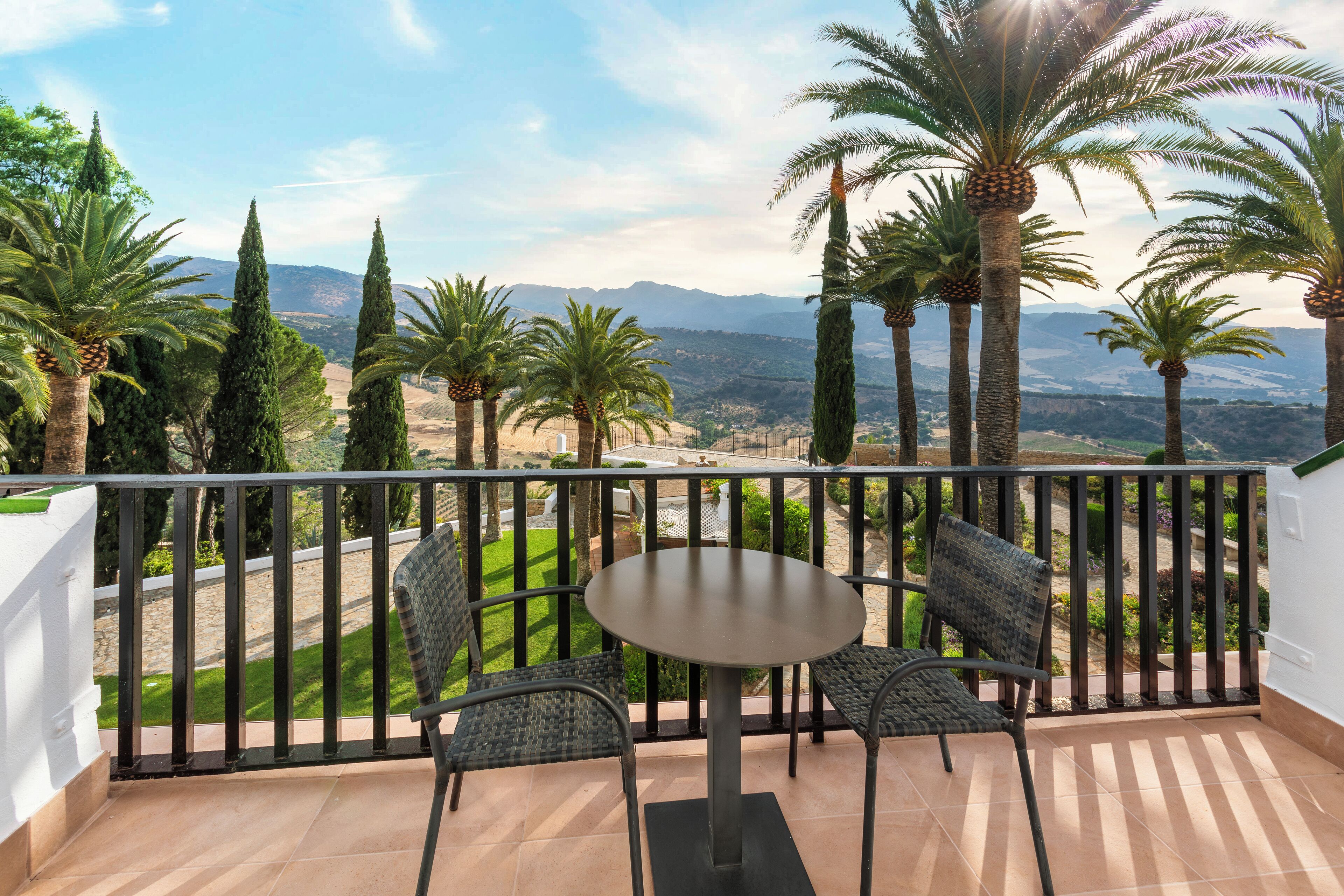 Ein Balkon mit rundem Tisch und zwei Stühlen, mit Blick auf eine malerische Berg- und Palmenlandschaft.