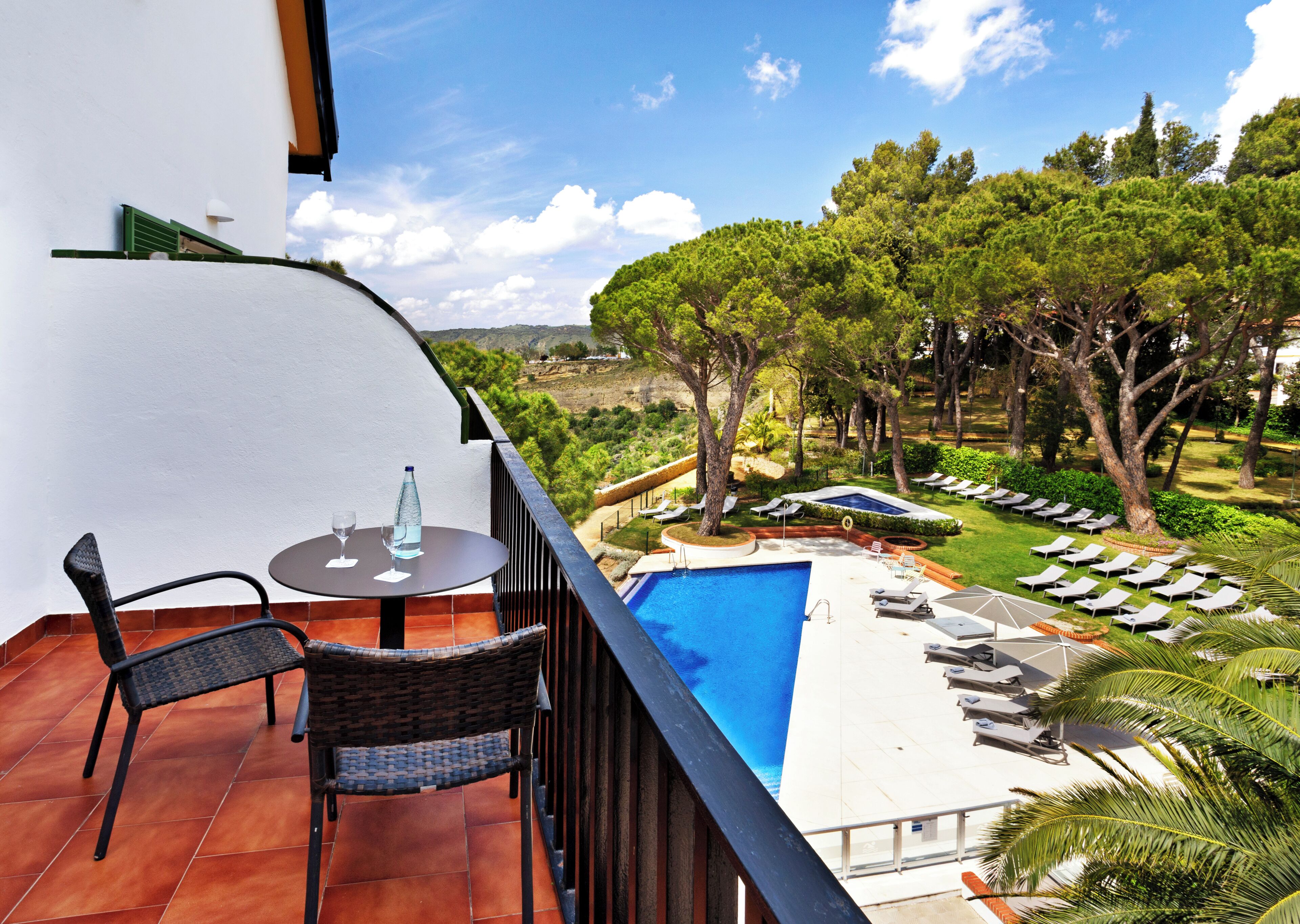 Un balcone con tavolo e sedie, affacciato su una piscina e un'area relax con alberi e vista sul paesaggio.
