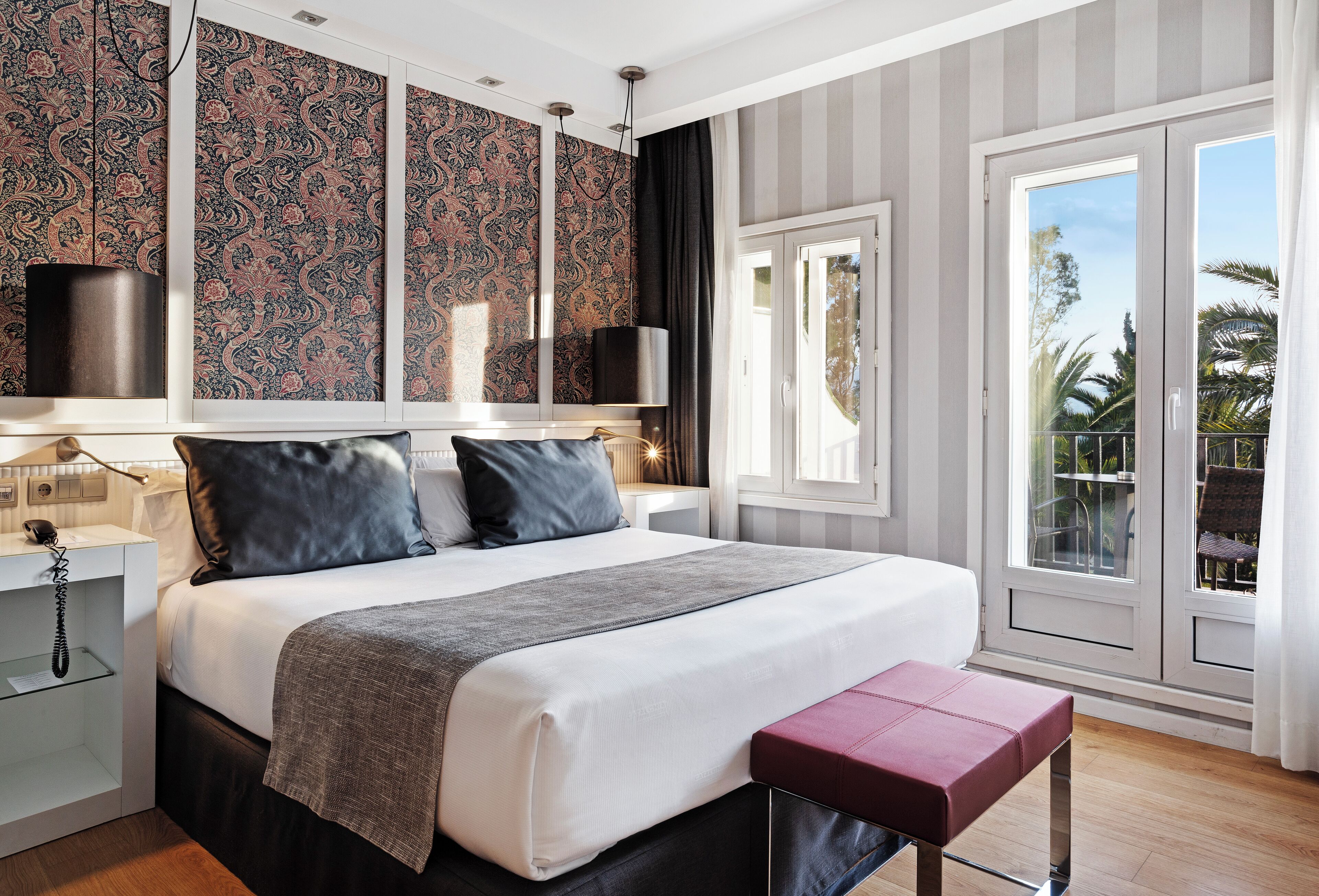 Habitación de hotel moderna con una cama grande, una mesita de noche con lámpara, un teléfono y vistas al exterior a través de una puerta corrediza de vidrio.
