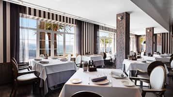 Un restaurante con mesas dispuestas para comer, amplias ventanas con cortinas y vistas a las montañas.