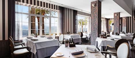 Un restaurante con mesas dispuestas para comer, amplias ventanas con cortinas y vistas a las montañas.