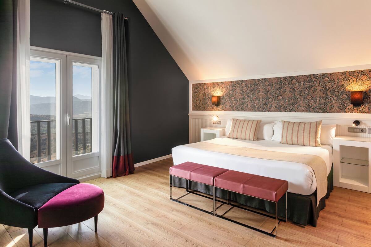 Ein Schlafzimmer mit einem großen Bett, einer Bank, einem Sessel und Blick ins Freie durch ein Fenster.