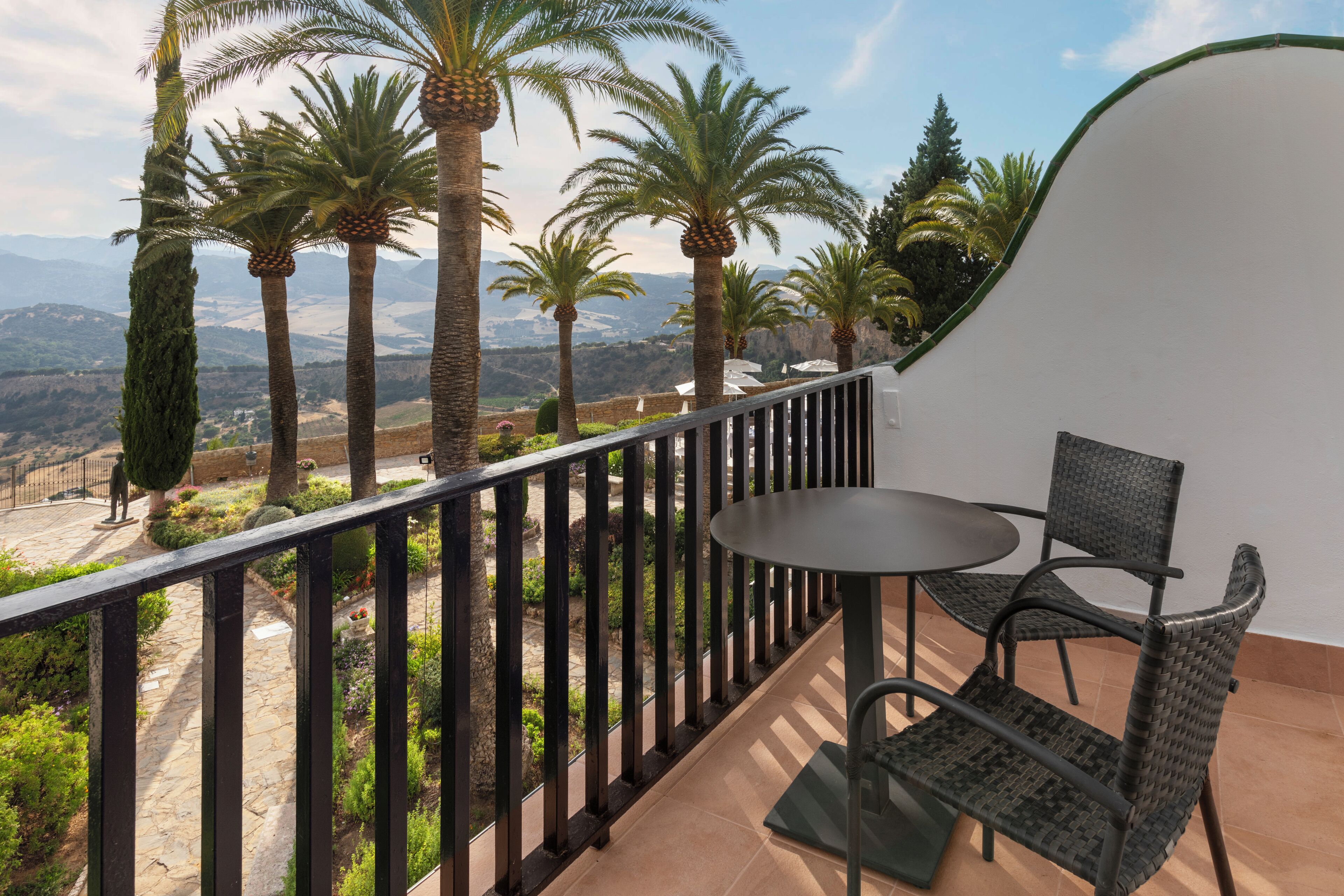 A balcony with a table and chairs, overlooking a scenic landscape with palm trees and mountains.