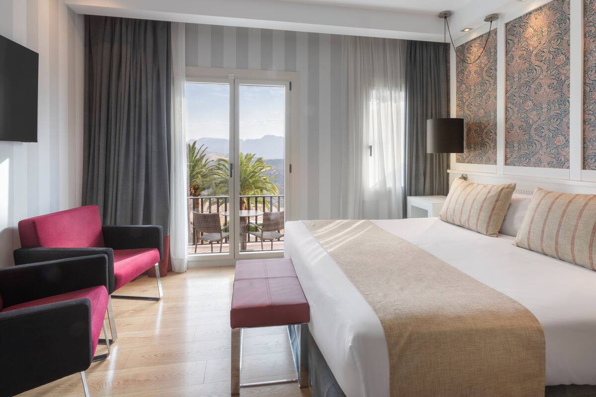 Ein Hotelzimmer mit einem großen Bett, einer Sitzecke mit Stühlen, einem Balkon mit Bergblick und einer Wand mit Muster.