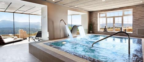 A modern indoor pool with a waterfall feature, large windows offering a mountain view, and a lounge area with seating.