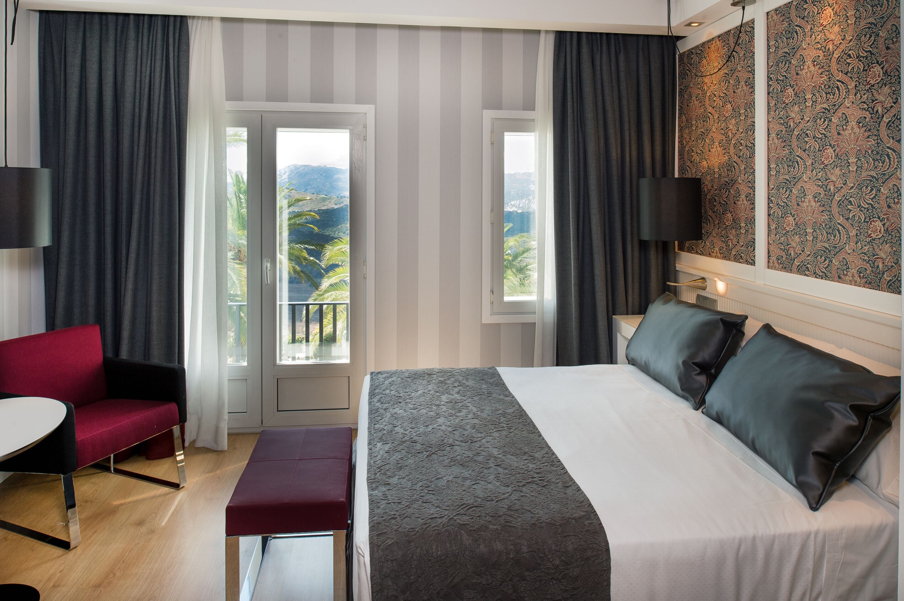 A hotel room with a bed, a desk, a red chair, and two windows offering a view of palm trees and mountains.