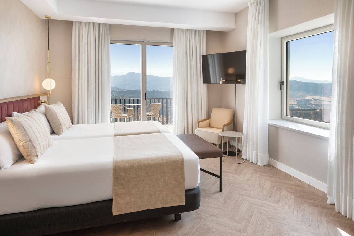 Ein Hotelzimmer mit einem großen Bett, einem Fernseher, einem Balkon mit Sitzgelegenheiten und Blick auf die Berge.